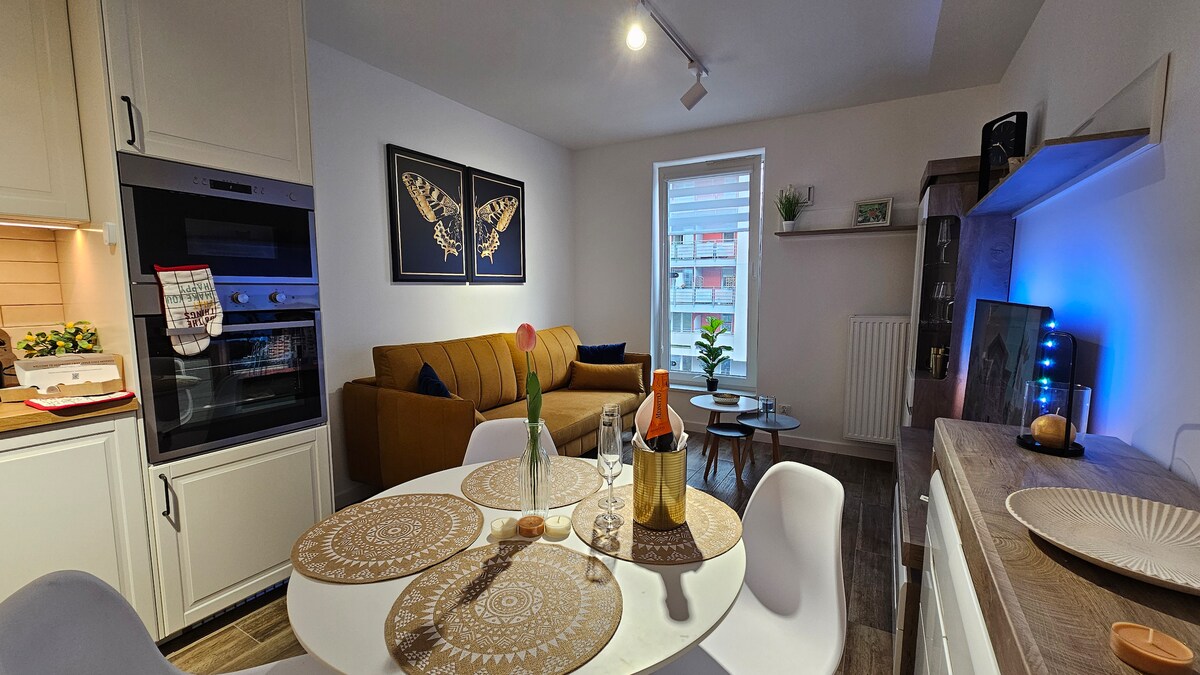 An inviting living area features a comfortable brown sofa and a round dining table set with decorative placemats. The modern kitchen is equipped with an oven and sleek cabinetry. Framed butterfly artwork adorns the walls, complementing indoor plants positioned by the window.