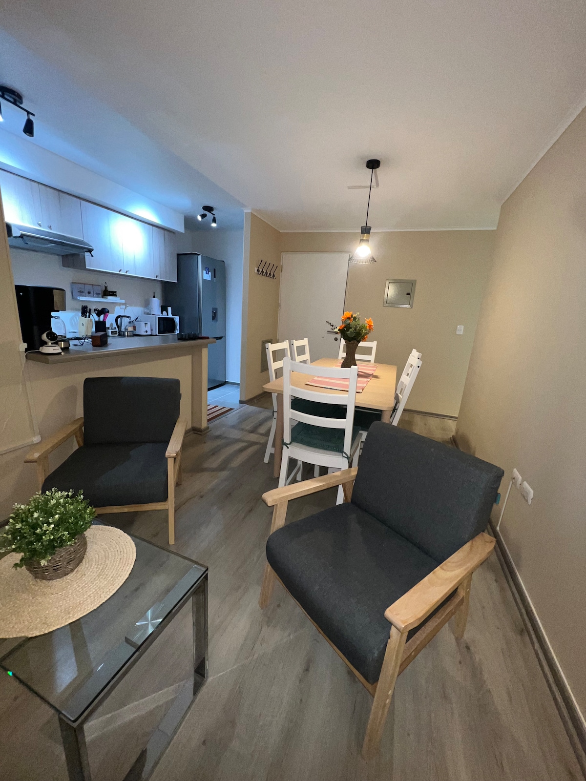 A living and dining area is presented, featuring a wooden table surrounded by white chairs and a flower centerpiece. Two grey armchairs are positioned nearby, and a glass-topped table holds a small potted plant. Soft lighting enhances the inviting interior design.