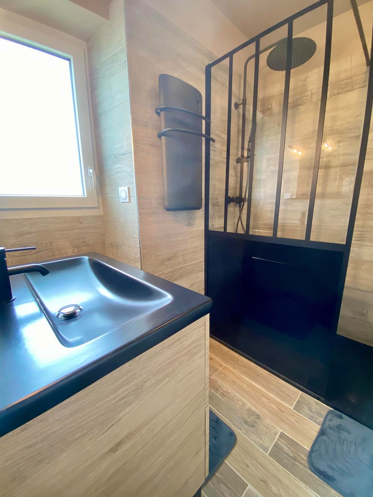 A modern bathroom features a sleek, dark sink and a glass shower enclosure with black metal accents. Natural light enters through a window, illuminating the textured wall tiles in neutral tones, creating a clean and functional space.