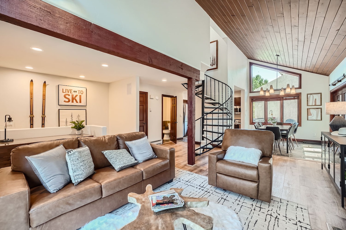 The living area features a brown leather sectional sofa paired with a comfortable armchair. A round coffee table sits atop a textured area rug. A decorative wall sign reading 'LIVE TO SKI' complements the rustic aesthetic, while large windows allow natural light to brighten the space.