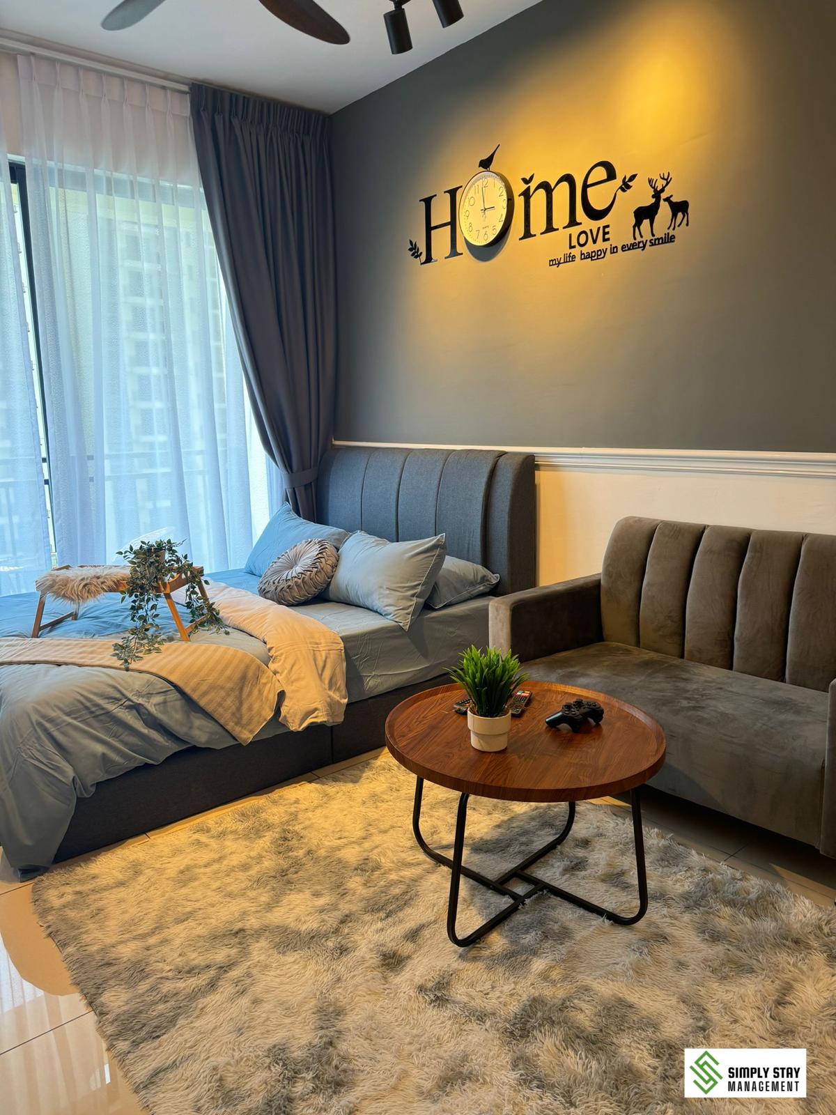 A cozy bedroom features a spacious bed with soft linens and decorative pillows. A sleek grey sofa is positioned beside it, offering additional seating. A round wooden coffee table rests on a soft area rug, which is complemented by a decorative plant. Large windows allow natural light to brighten the space.