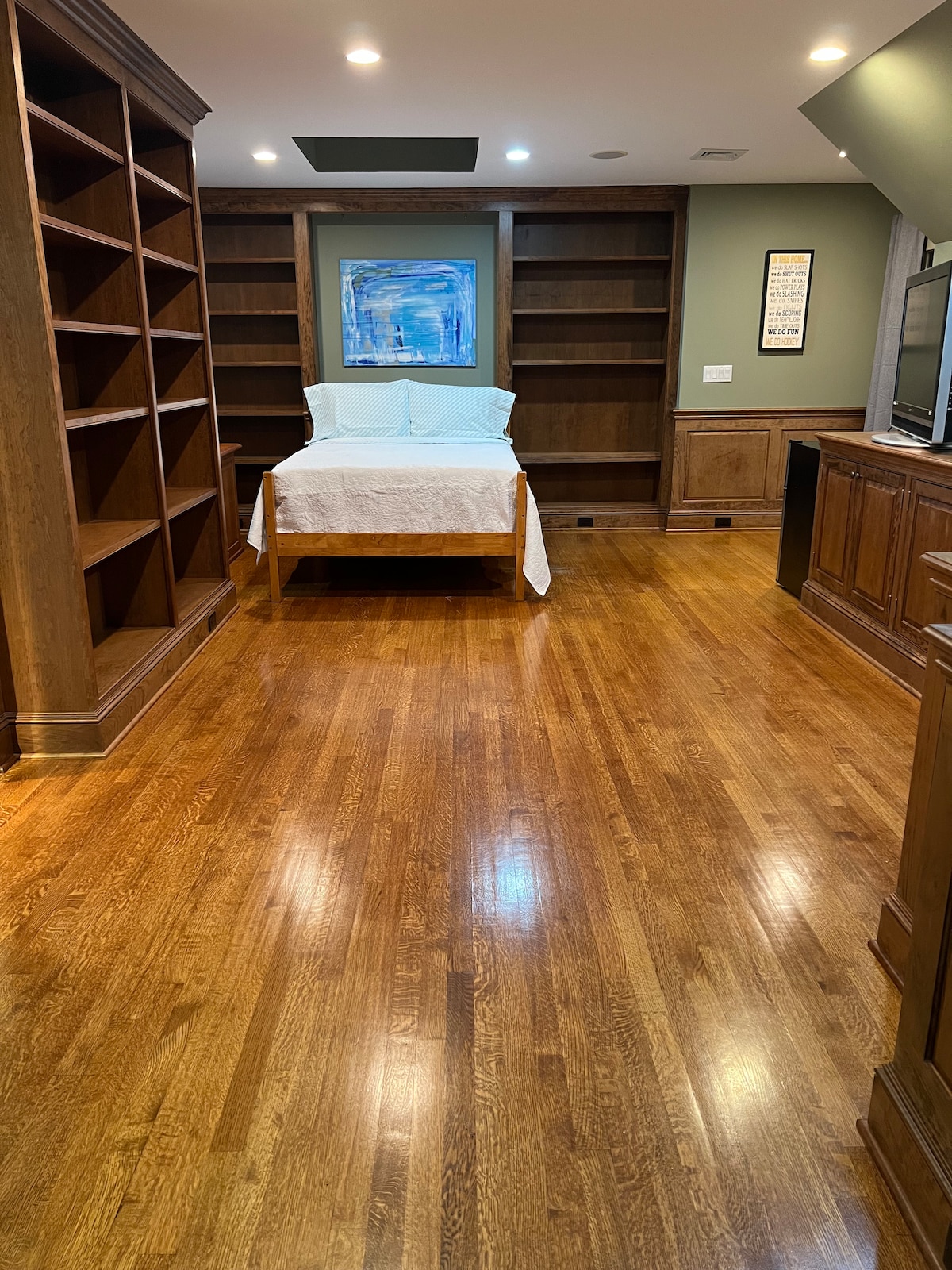 A spacious room features polished hardwood floors, complemented by built-in wooden shelves. A bed with a white coverlet occupies the center, and natural light brightens the space. A framed artwork adds a touch of color to the neutral walls, while modern lighting enhances the overall ambiance.