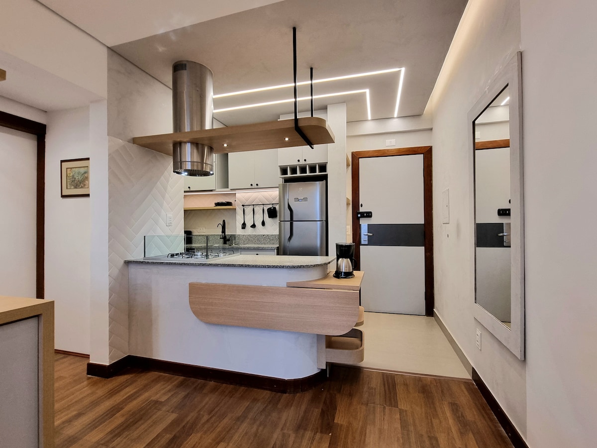 A modern kitchen area features a sleek design with light wood accents and stainless steel appliances. A central island provides additional cooking space, while overhead lighting enhances visibility. The entrance doorway is visible, framed by a reflective mirror, contributing to an open feel.