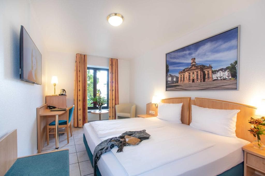 A well-lit room with two spacious beds is featured, accompanied by light-colored wooden furnishings. A flat-screen TV is mounted on the wall above a small desk and chair. Natural light enters through a window with curtains, offering views of the outside.