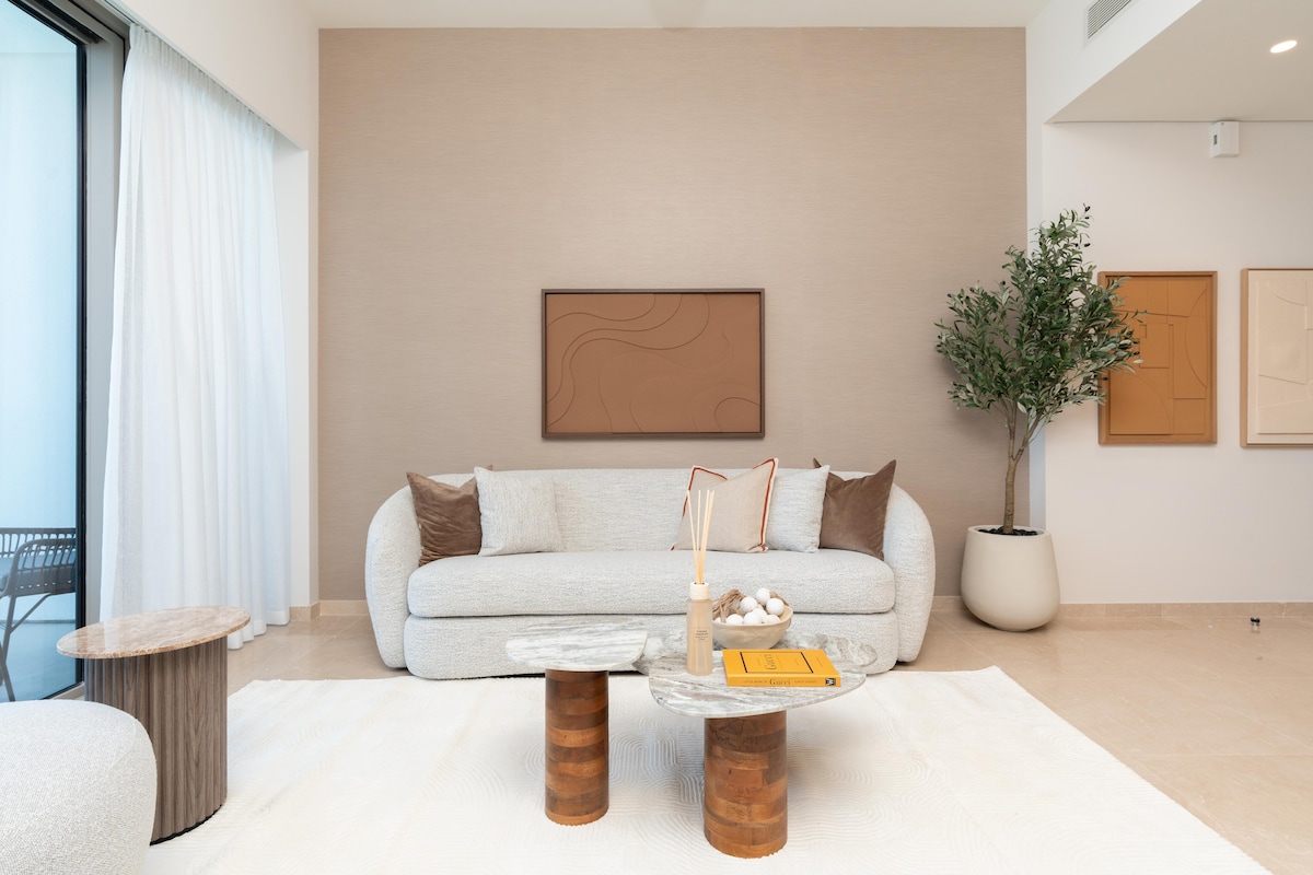 A spacious living area features a light-colored, curved sofa accompanied by two decorative pillows. A round coffee table made of marble and wood sits in front of the sofa, while a small side table complements the space. Natural light flows through floor-to-ceiling windows.