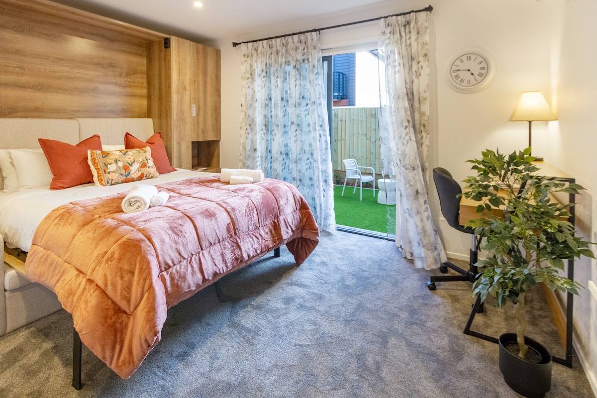 A cozy sleeping area is presented with a plush queen bed adorned in soft, warm bedding. Light curtains frame a glass door, leading to a private outdoor space. A compact workspace with a desk and chair, alongside a potted plant, complements the inviting room decor.