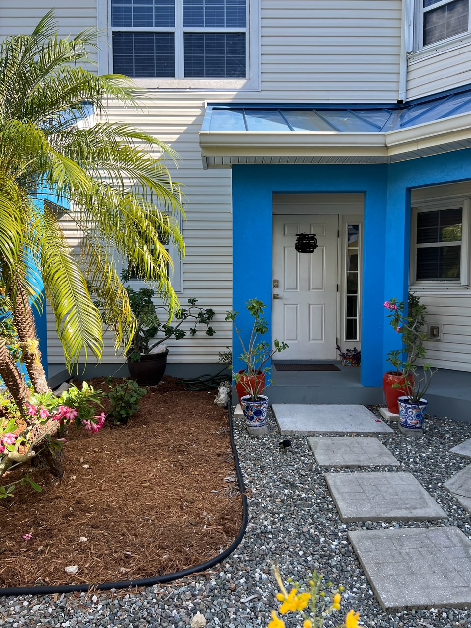 The exterior entrance features a vibrant blue trim surrounding the front door. A pathway of concrete stones leads through a gravel area, bordered by plants and flowers. Lush greenery, including a tall palm, adds a tropical touch to the inviting entryway.