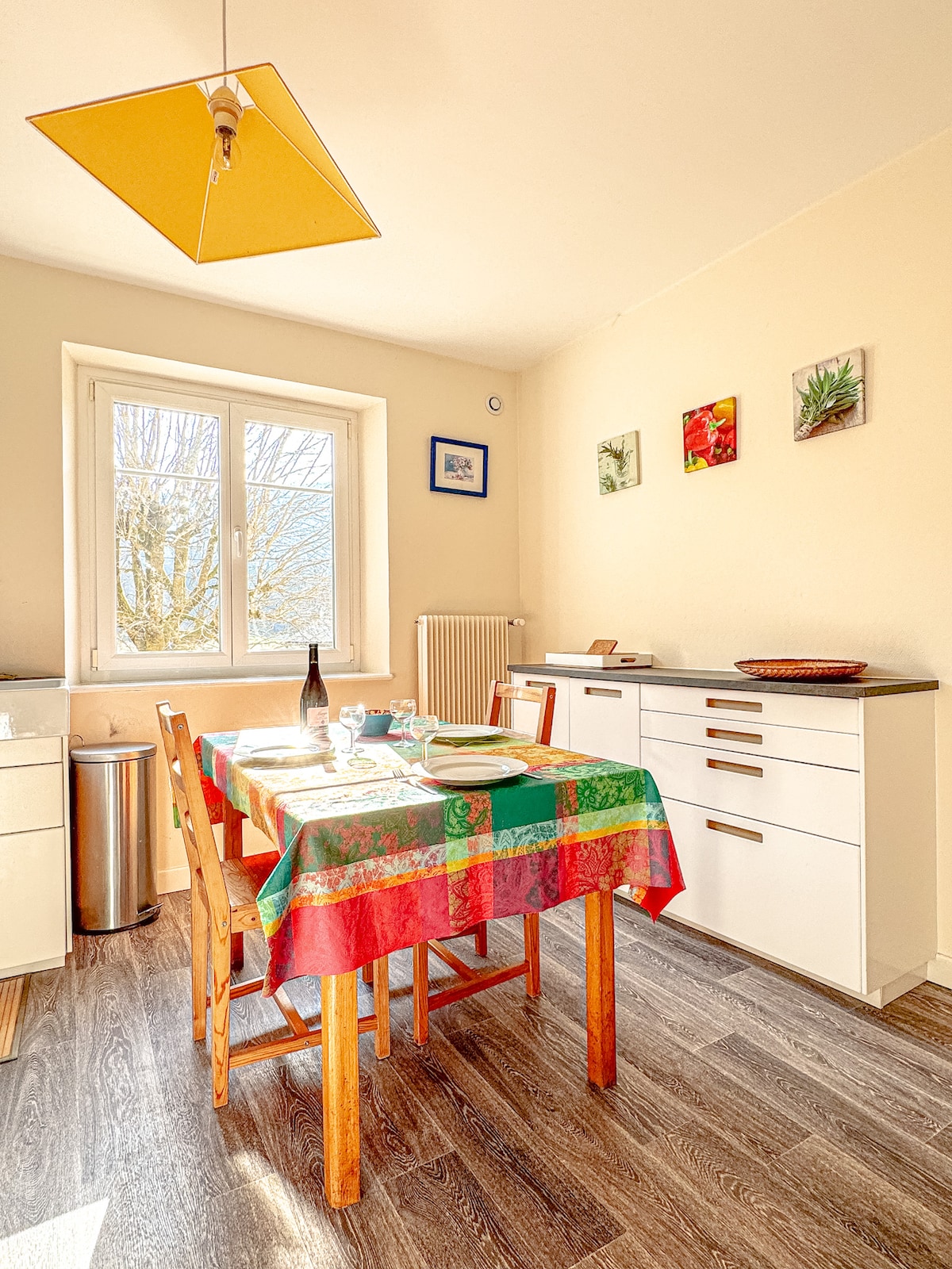 A dining area is presented with a colorful tablecloth draped over a wooden table, set with dishes and glassware. Light enters through a large window, illuminating the space. The walls display small, framed artwork, complementing the modern white cabinetry.