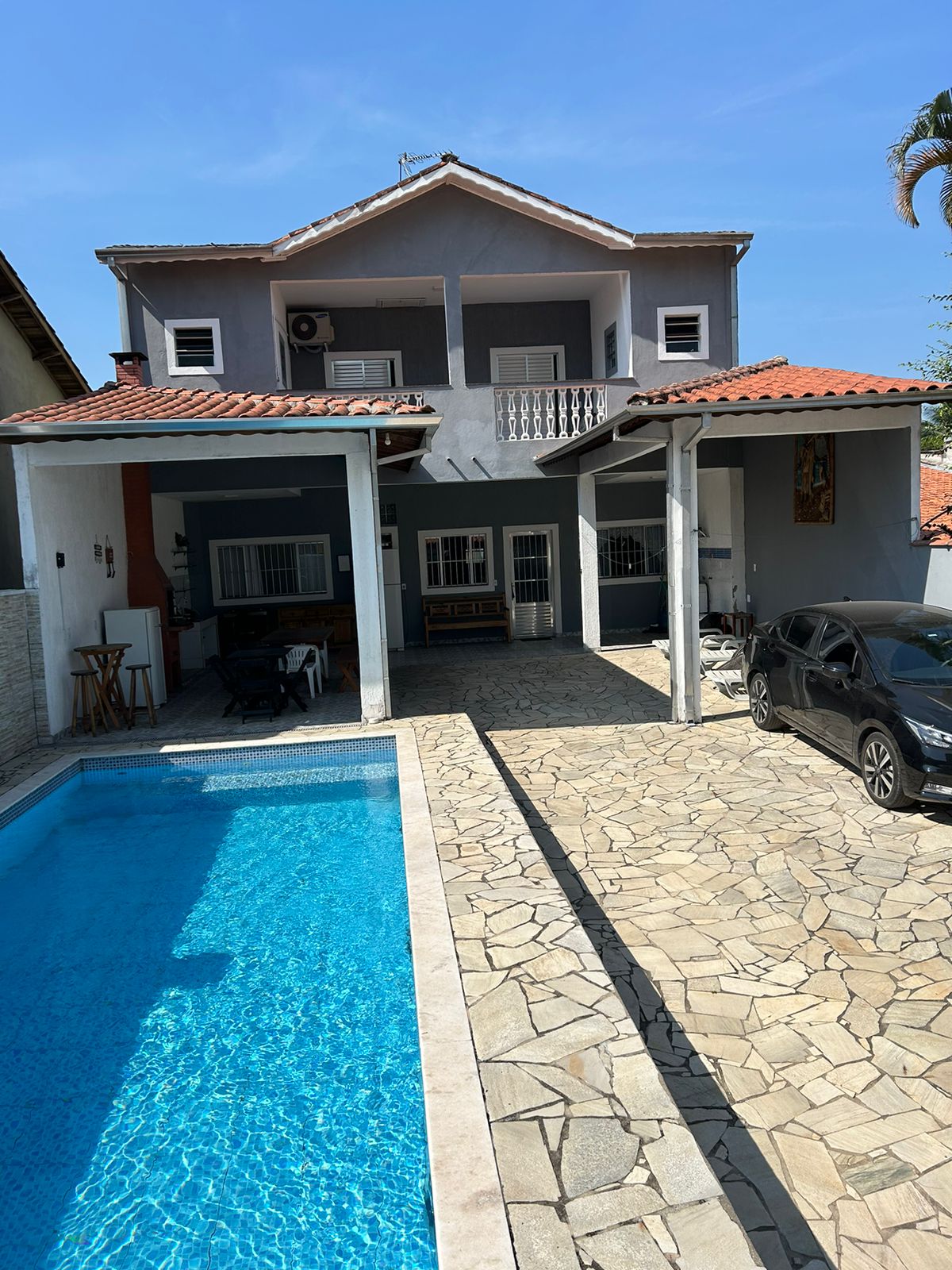 A modern, two-story house with a terracotta roof is showcased beside a refreshing pool. The outdoor area is paved with light stone, featuring ample space for relaxation. A shaded porch is visible, providing an inviting entrance to the home, while a parked car adds convenience to the setting.