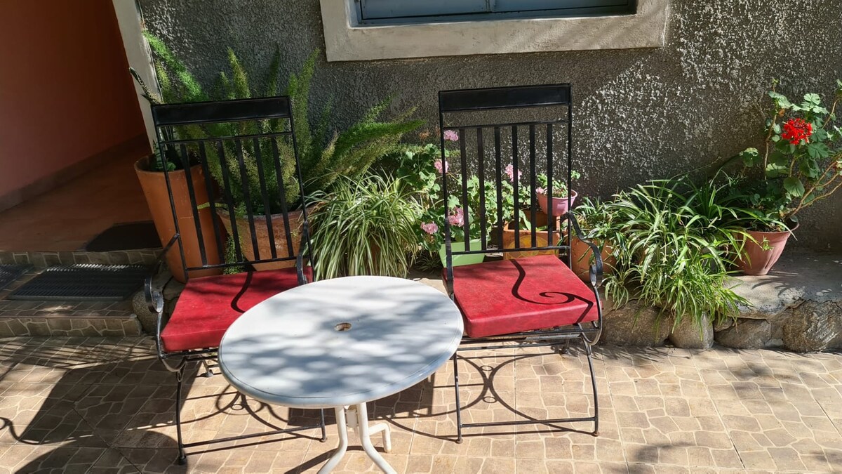 An outdoor seating area is displayed with two black chairs featuring red cushions positioned around a small round table. Potted plants and flowers add greenery to the space, contributing to a welcoming environment.