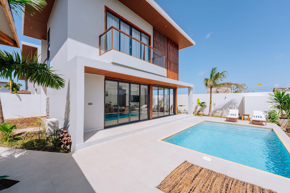 The exterior showcases a modern villa with large glass doors that create a seamless connection between indoor and outdoor spaces. A private pool is surrounded by lush greenery and loungers, while a natural fiber rug adds warmth to the patio area.