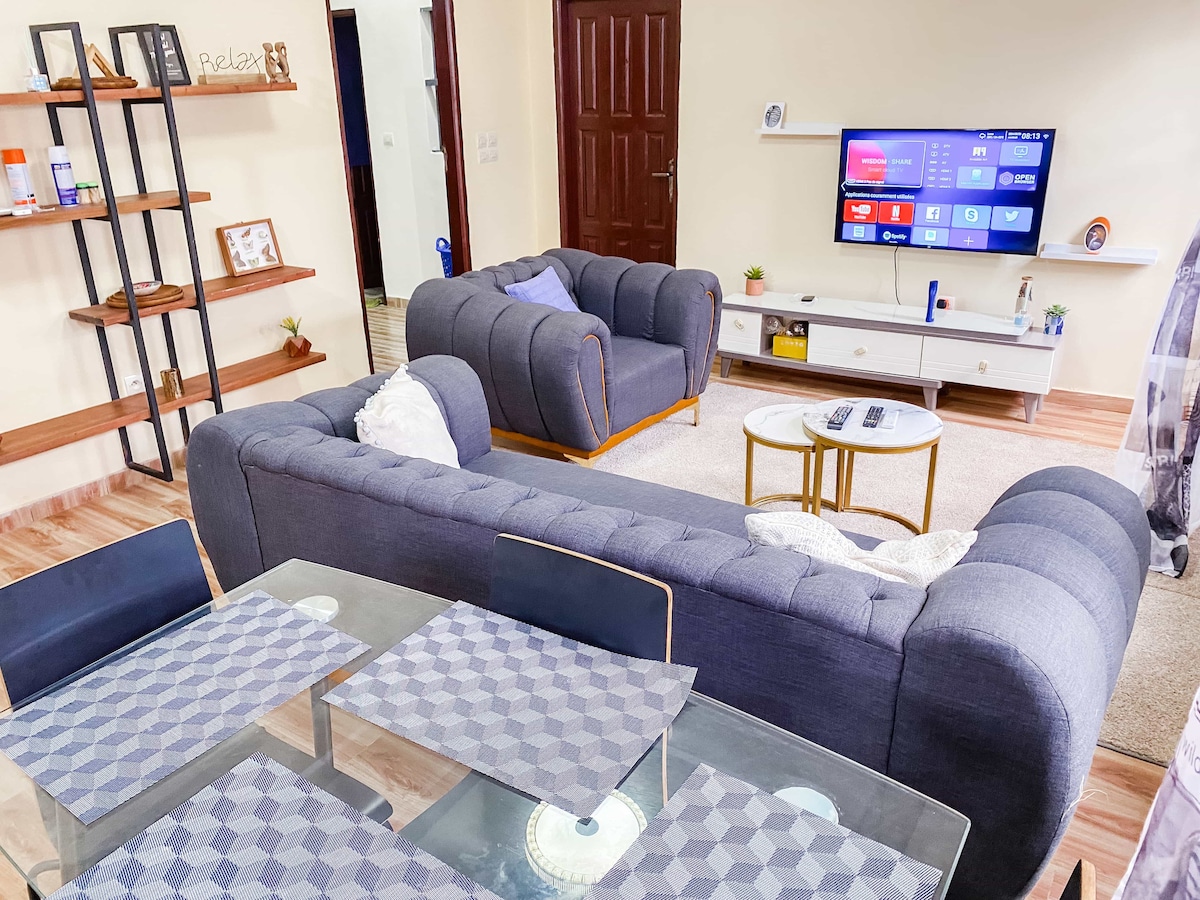 A cozy living room is presented with comfortable gray sofas and a glass coffee table. A television is mounted on the wall, providing entertainment options. Decorative shelves hold various items, contributing to the room's inviting ambiance. Light wood flooring complements the overall design.