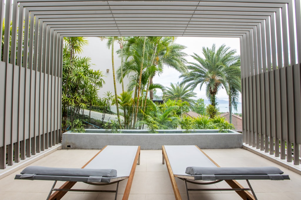 A serene relaxation area is presented, featuring two lounge chairs positioned on a light-colored tiled surface. Lush greenery surrounds the space, with tall palm trees visible through the linear slatted structure that frames the view.