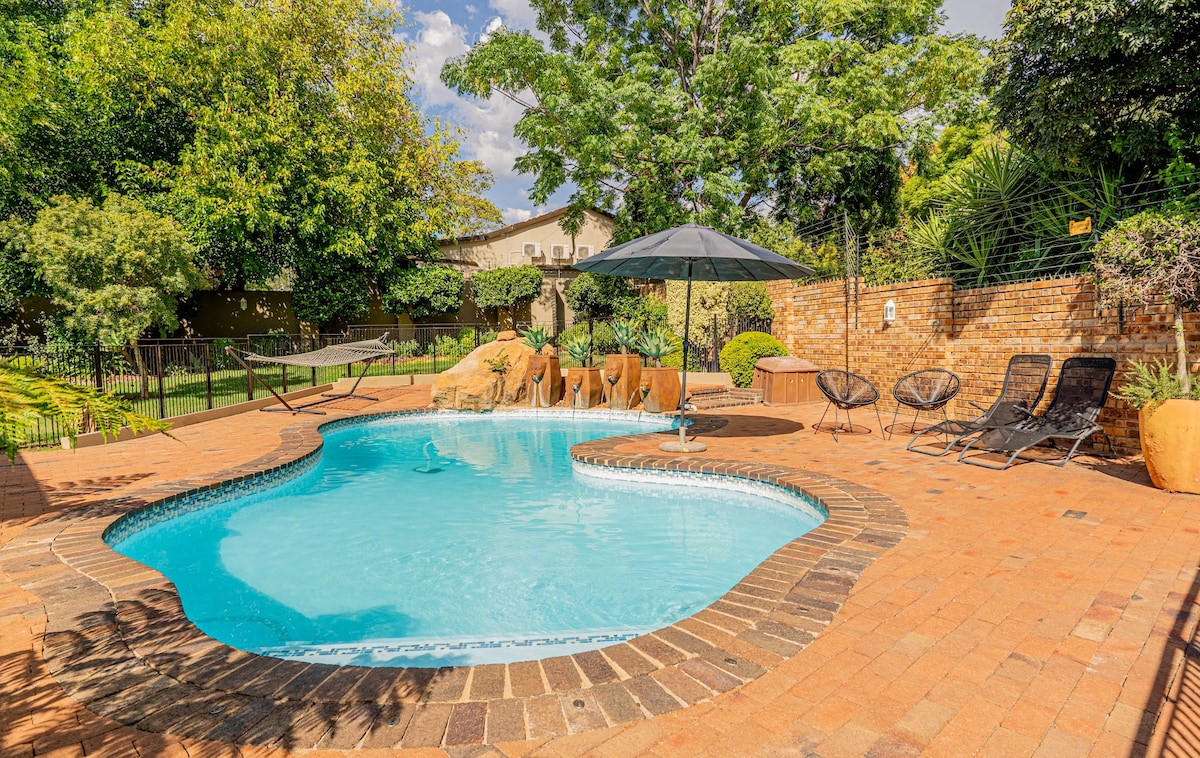 A seasonal swimming pool is surrounded by a spacious patio, featuring comfortable lounge chairs and an umbrella for shade. Lush greenery lines the perimeter, and a hammock is visible nearby, offering a serene outdoor space for relaxation.