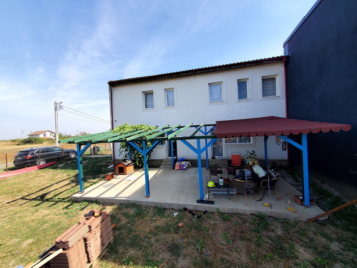 The spacious outdoor area features a covered patio supported by blue beams, providing shade over various seating arrangements. A small structure is visible in the corner, surrounded by grass, with the home’s white exterior and multiple windows in the background.