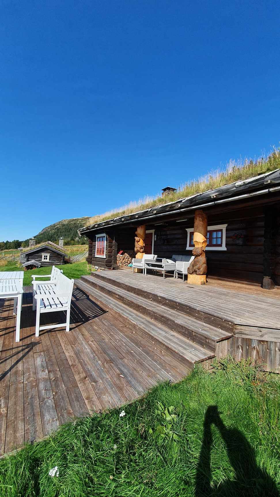 A wooden cabin exterior is visible under a clear blue sky. The spacious deck features wooden flooring and traditional sculptures, complemented by white outdoor furniture. Lush grass surrounds the area, enhancing the natural setting of the cabin.