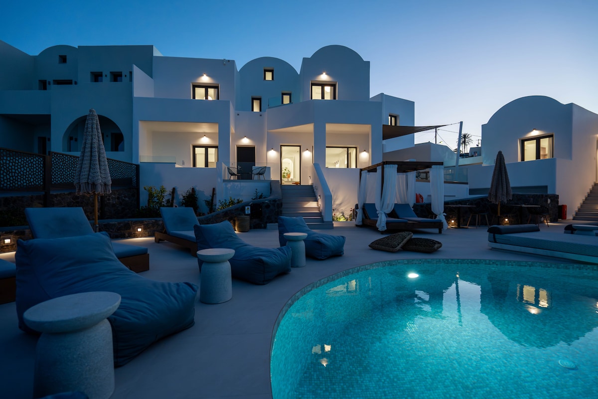 An outdoor area is featured with a illuminated swimming pool reflecting soft blue hues. Lounge chairs are positioned around the pool, with some shaded by large umbrellas. White-walled buildings, designed with rounded edges, are visible in the background against the twilight sky.