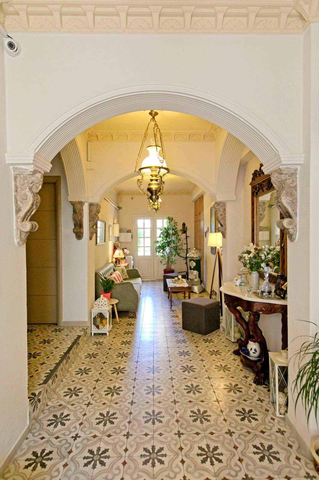 A bright hallway is framed by an ornate archway, featuring traditional tile flooring with intricate patterns. Soft lighting is provided by pendant and table lamps, illuminating a cozy seating area to the left. Decorative plants and wall art enhance the welcoming ambiance.