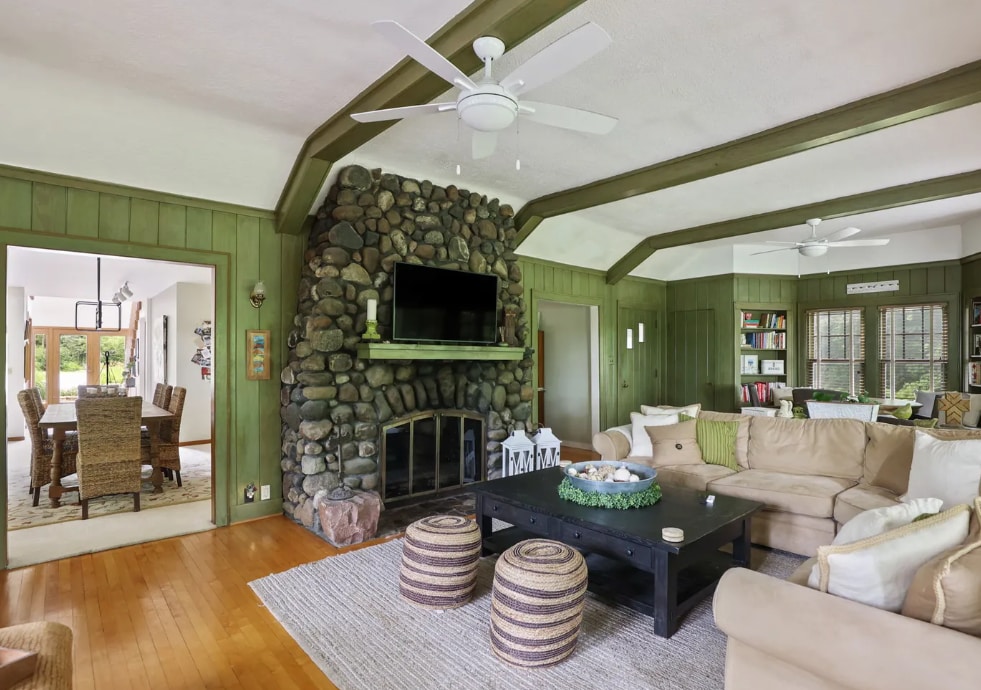 A spacious great room features a stone fireplace and a flat-screen television. Natural light is welcomed through large windows, highlighting green waxed wood paneling and original wood flooring. Comfortable seating arrangements are provided with soft couches and decorative poufs, creating an inviting gathering space.
