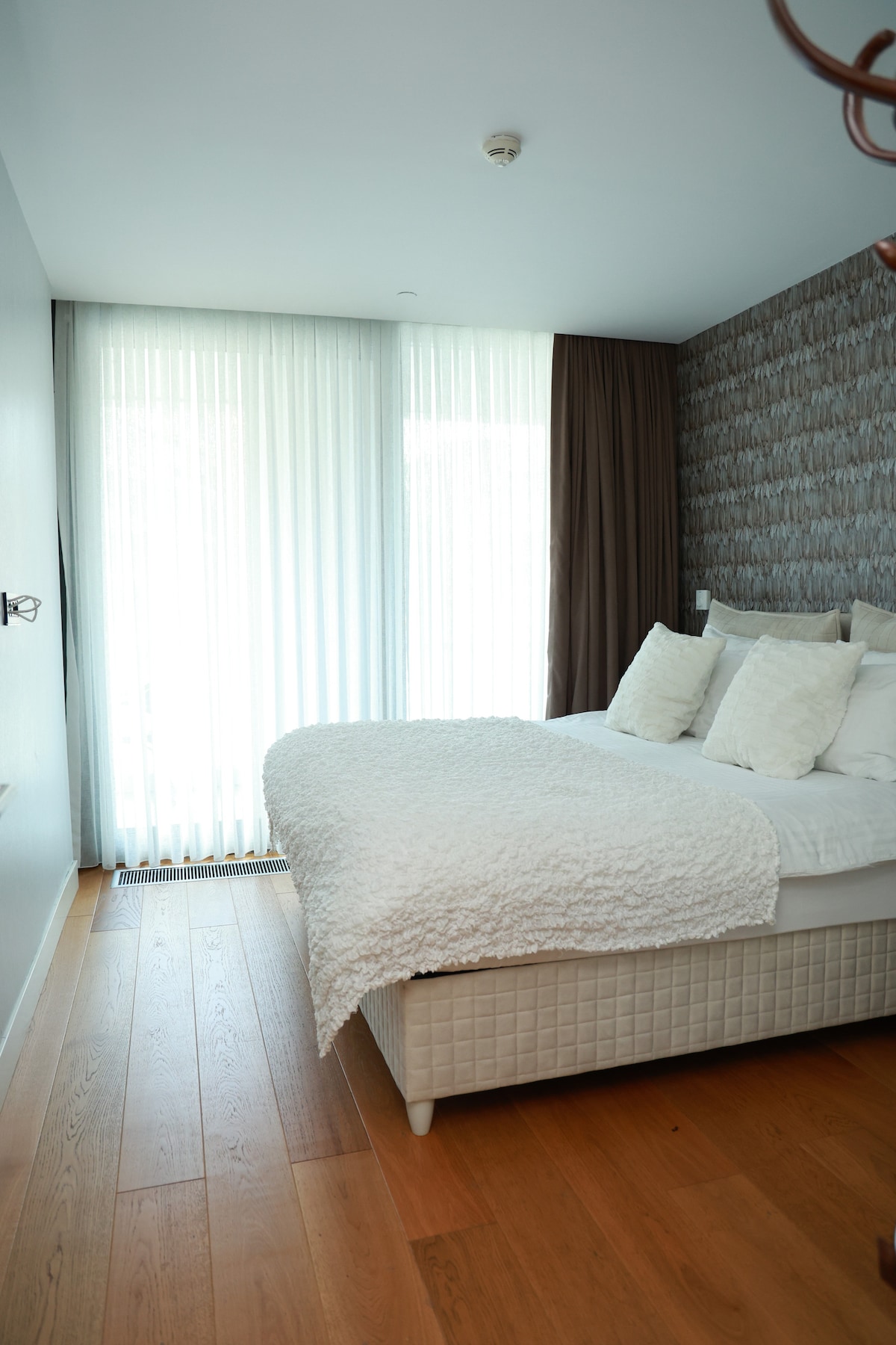 A well-appointed bedroom is presented with a spacious bed layered with a textured white blanket and decorative pillows. Floor-to-ceiling sheer curtains allow natural light to fill the room, highlighting the light wood flooring.