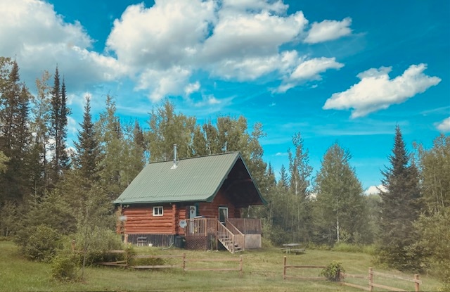 The rustic cabin, featuring a sloped green roof and wooden exterior, is surrounded by lush trees and open grass. A wooden deck leads to the entrance, with a picnic table visible nearby. The clear blue sky is dotted with fluffy white clouds.