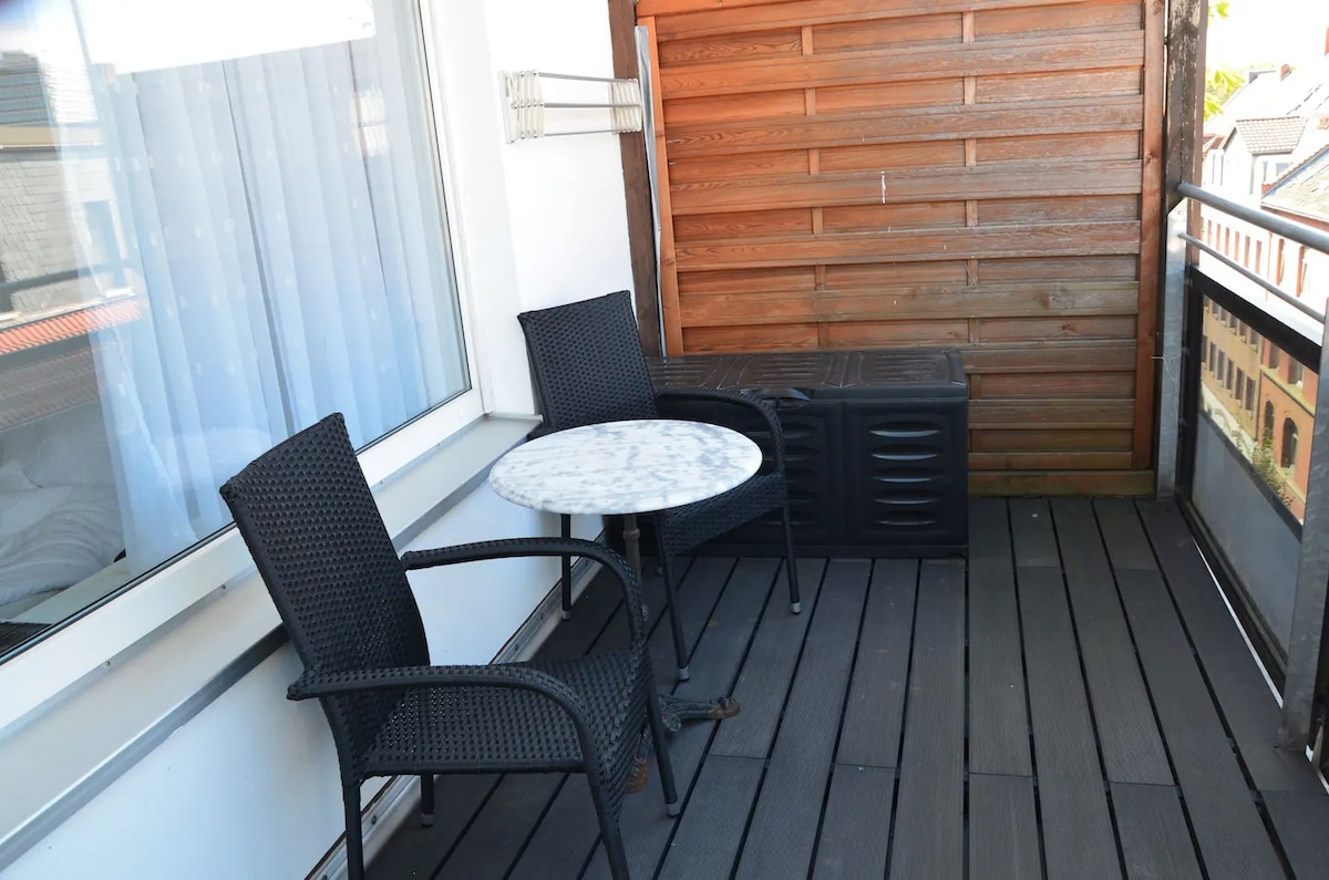 A small balcony features a round marble table flanked by two black wicker chairs. A storage chest is positioned against a wooden wall, while large windows allow light to filter in from the interior space.