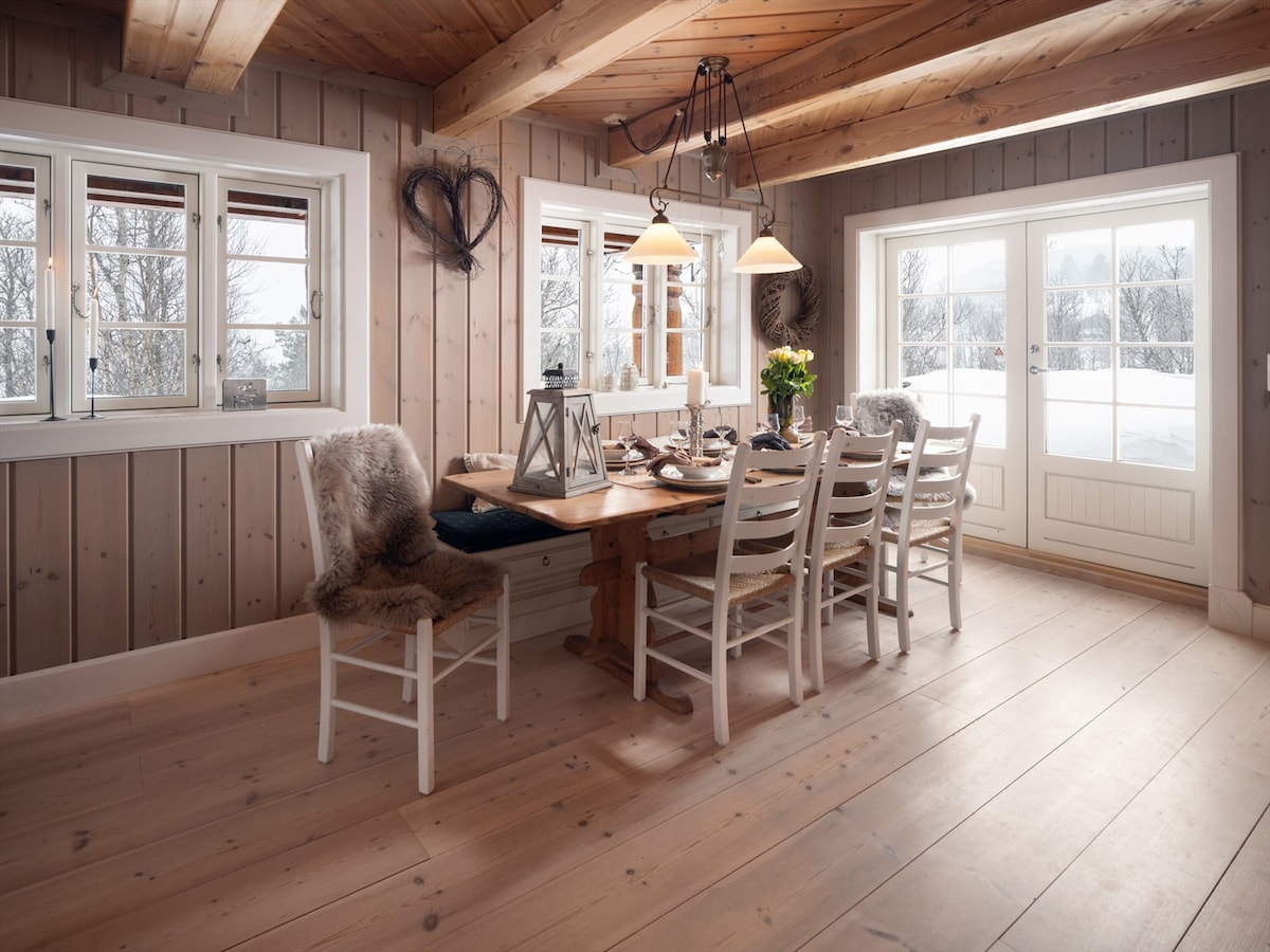 A dining area featuring a large wooden table surrounded by six white chairs. Two pendant lights hang above the table. The windows allow natural light to fill the room, showcasing a snowy landscape outside. The wooden walls and ceiling create a rustic atmosphere.