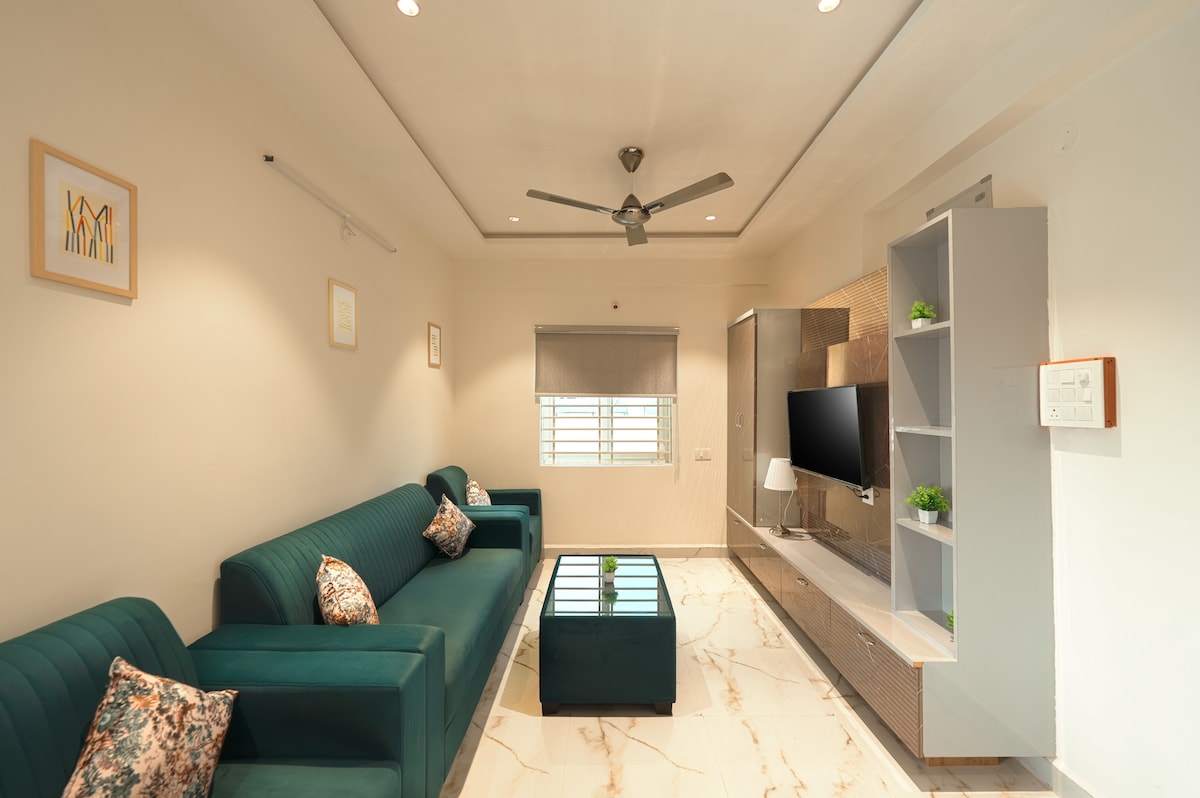 A modern living area features a teal sofa and a matching ottoman set against a light-toned wall. A flat-screen TV is mounted on a stylish unit, complemented by decorative shelving. Natural light enters through a window, and a ceiling fan adds functionality.