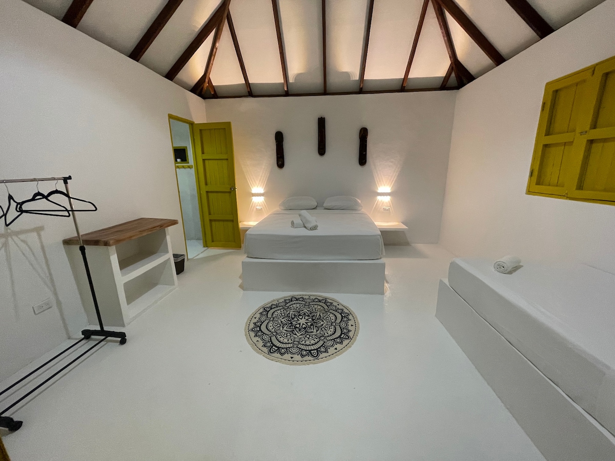 A bright room features a spacious bed adorned with crisp white linens. Two bedside lamps gently illuminate the space. A decorative rug adds texture to the polished floor. Yellow window shutters and wall accents provide a cheerful contrast, while a clothing rack and small table complete the layout.