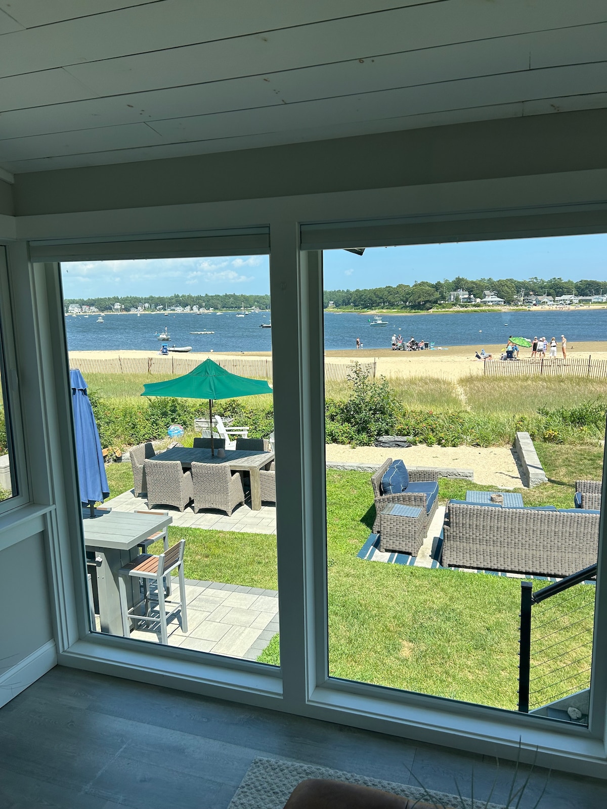 A view from inside showcases large glass windows framing a serene scene of the beach and waterfront. Outdoor seating and tables are visible on the patio, with individuals enjoying leisure time on the sandy shore across the water.