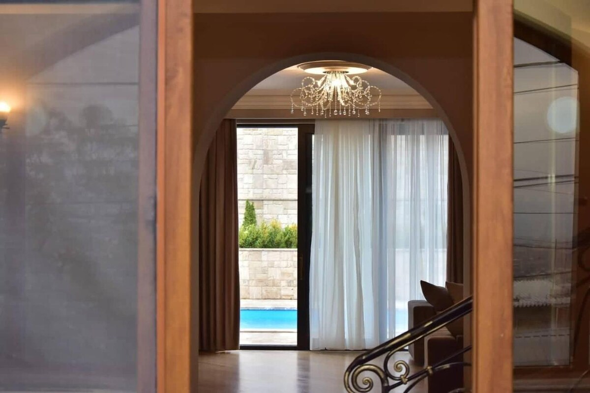 An elegant entryway features an arched transition, framed by light curtains that gently sway. A chandelier hangs from the ceiling, illuminating the space. Through the glass doors, the serene outdoor pool area is partially visible, enhancing the inviting and calm atmosphere.