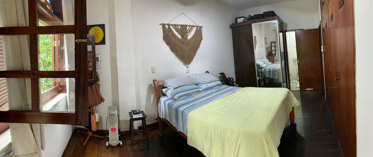 A spacious bedroom features a large bed with blue and cream striped bedding and natural light filtering through wooden shutters. A macramé wall hanging is displayed above the bed. A wooden wardrobe and side tables complement the dark wooden flooring, creating a cozy atmosphere.