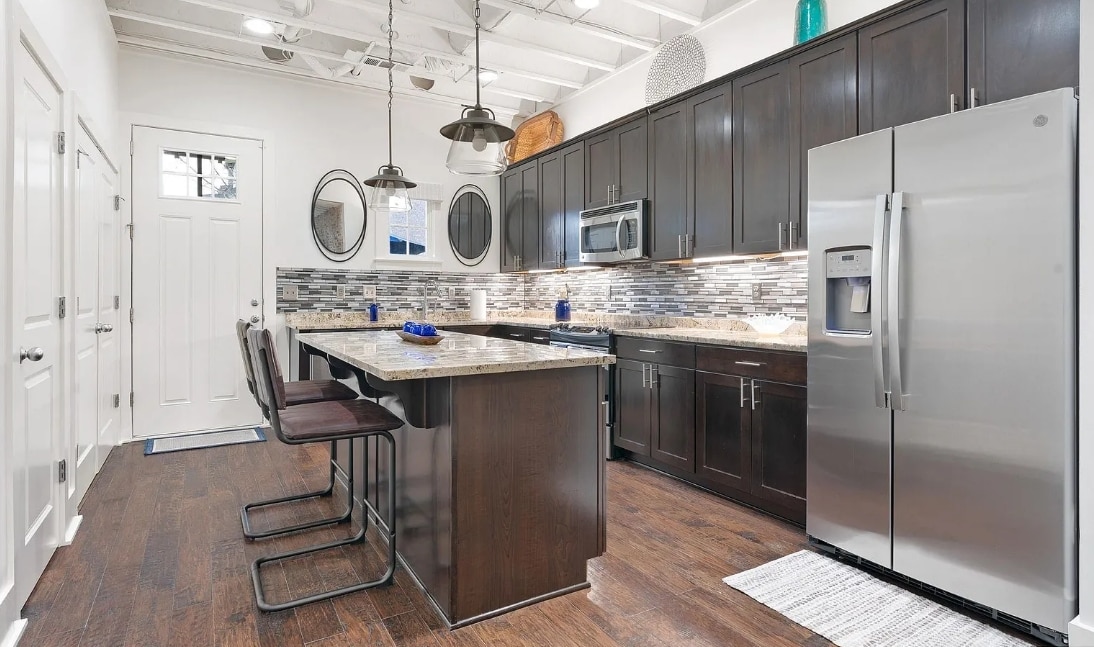 The kitchen showcases dark wooden cabinetry and a large island with a granite countertop. Stainless steel appliances, including a refrigerator, are visible. Two bar stools are positioned at the island. Natural light filters in through a nearby door, highlighting the modern tile backsplash.