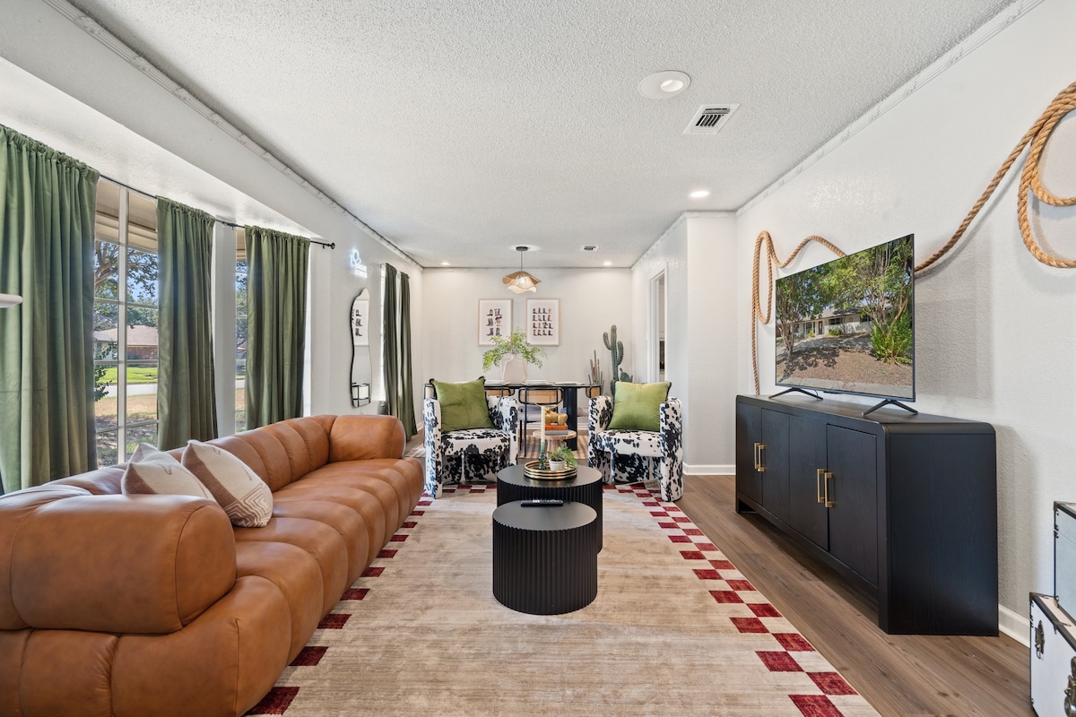 A spacious living room showcases a large brown couch paired with thematic chairs, providing ample seating. A coffee table is centered on a light area rug, while a smart TV is positioned against a sleek black cabinet. Floor-to-ceiling windows with green curtains allow natural light to enter.