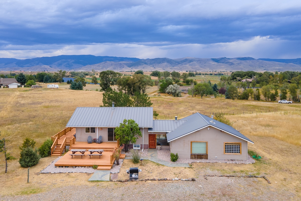 A charming retreat is set against a backdrop of rolling hills and the Absaroka Mountains. The home features a mix of wood and light-colored siding, surrounded by grassy expanses. An inviting deck with picnic tables provides a space for outdoor gatherings, complemented by the natural landscape.