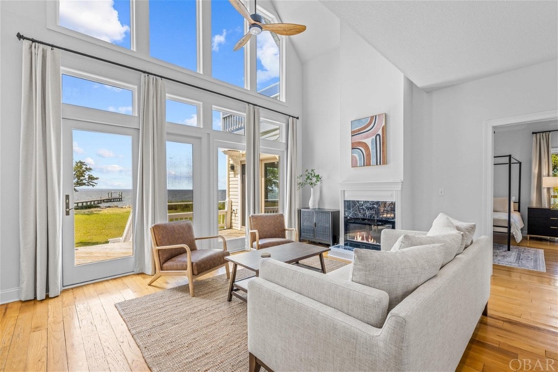 A spacious living area features large windows that offer views of the sound. A comfortable light-colored sofa and two wooden chairs are arranged around a sleek coffee table. Natural light floods the room, enhancing the clean, modern aesthetic complemented by a fireplace and decorative wall art.