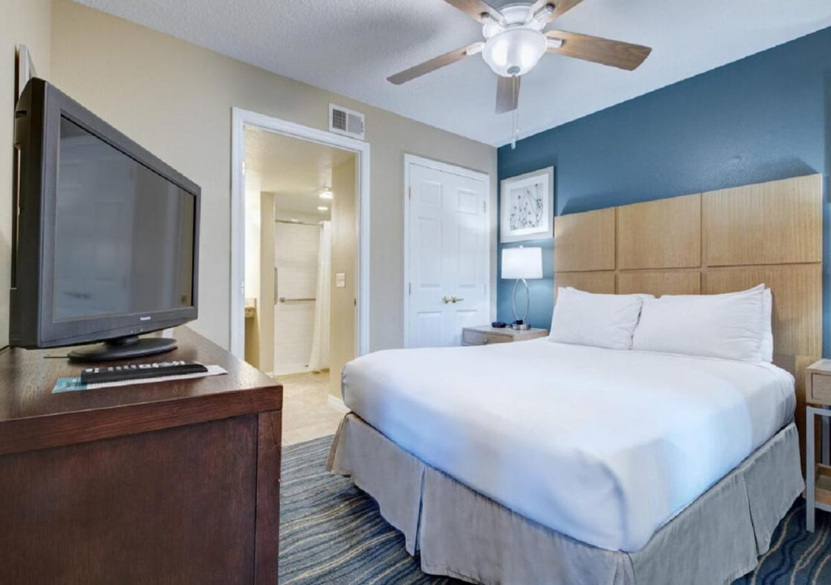 A well-lit bedroom features a spacious bed with white linens and a decorative headboard. A television is positioned on a wooden dresser beside the bed. A ceiling fan is mounted above, and a bathroom door is visible in the background.