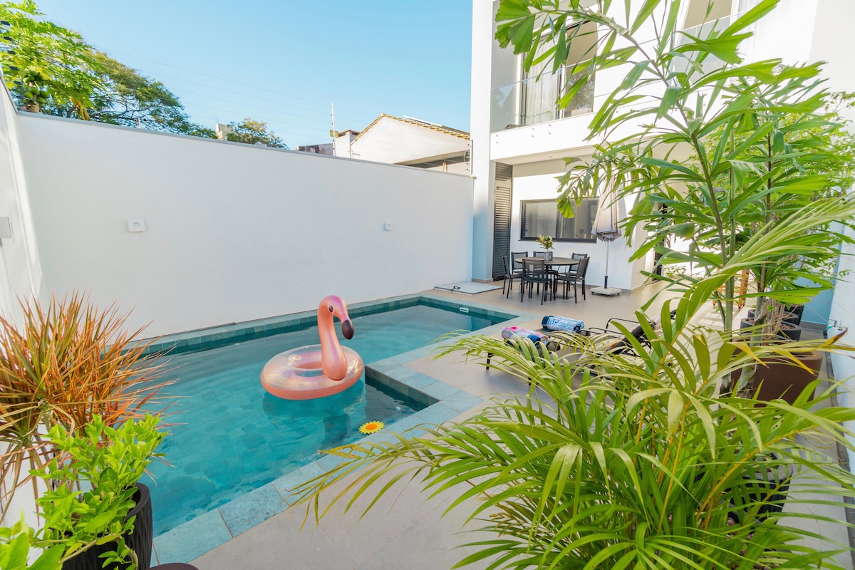 A private pool area is surrounded by greenery, featuring a bright inflatable flamingo floating on the water. Lounge chairs are positioned nearby, with a dining table set under a shaded area for six guests. Natural light enhances the tranquil atmosphere.