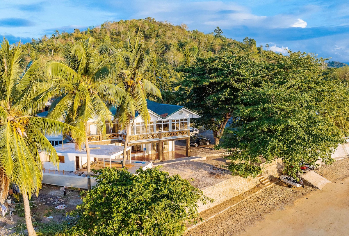 A coastal property is set against a verdant hillside, featuring palm trees and spacious outdoor areas. The building showcases a wraparound balcony and large windows, inviting natural light into the interior. The surrounding landscape highlights a serene beach environment.