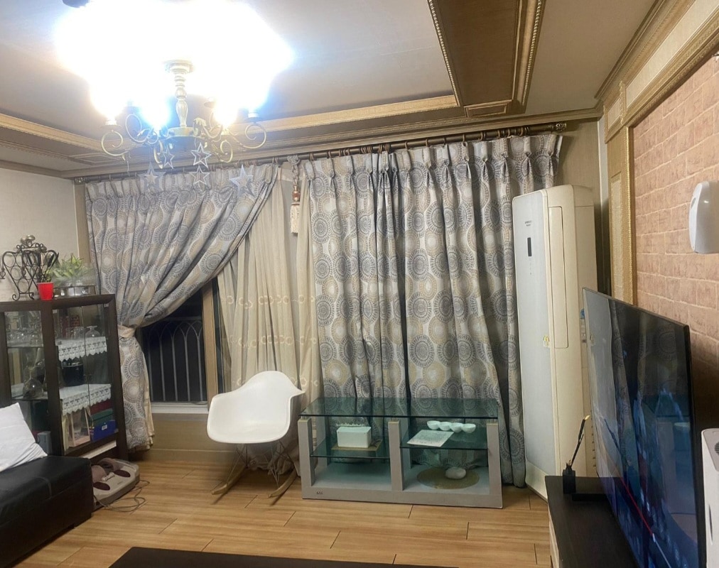 A living area features a stylish chandelier hanging from the ceiling. Elegant curtains frame a nearby window, while a contemporary chair is positioned next to a glass coffee table. A television sits on a stand, complemented by a display cabinet filled with various items.