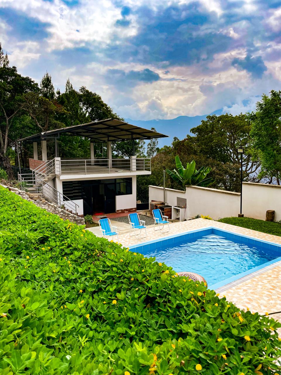 A well-maintained garden is displayed in the foreground, leading to a sparkling pool surrounded by a patio. Near the pool, lounge chairs are arranged, offering a space for relaxation. A modern building with large windows is visible, providing a connection to the natural surroundings.
