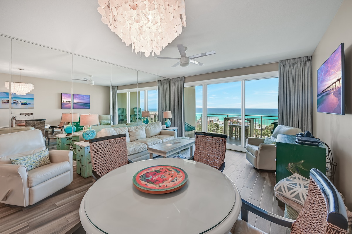 A spacious living area features a light-colored sofa set and a round dining table with textured chairs. Large glass doors offer expansive ocean views, while decorative elements, such as unique lighting fixtures and coastal decor, enhance the atmosphere. Mirrored walls reflect the natural light.