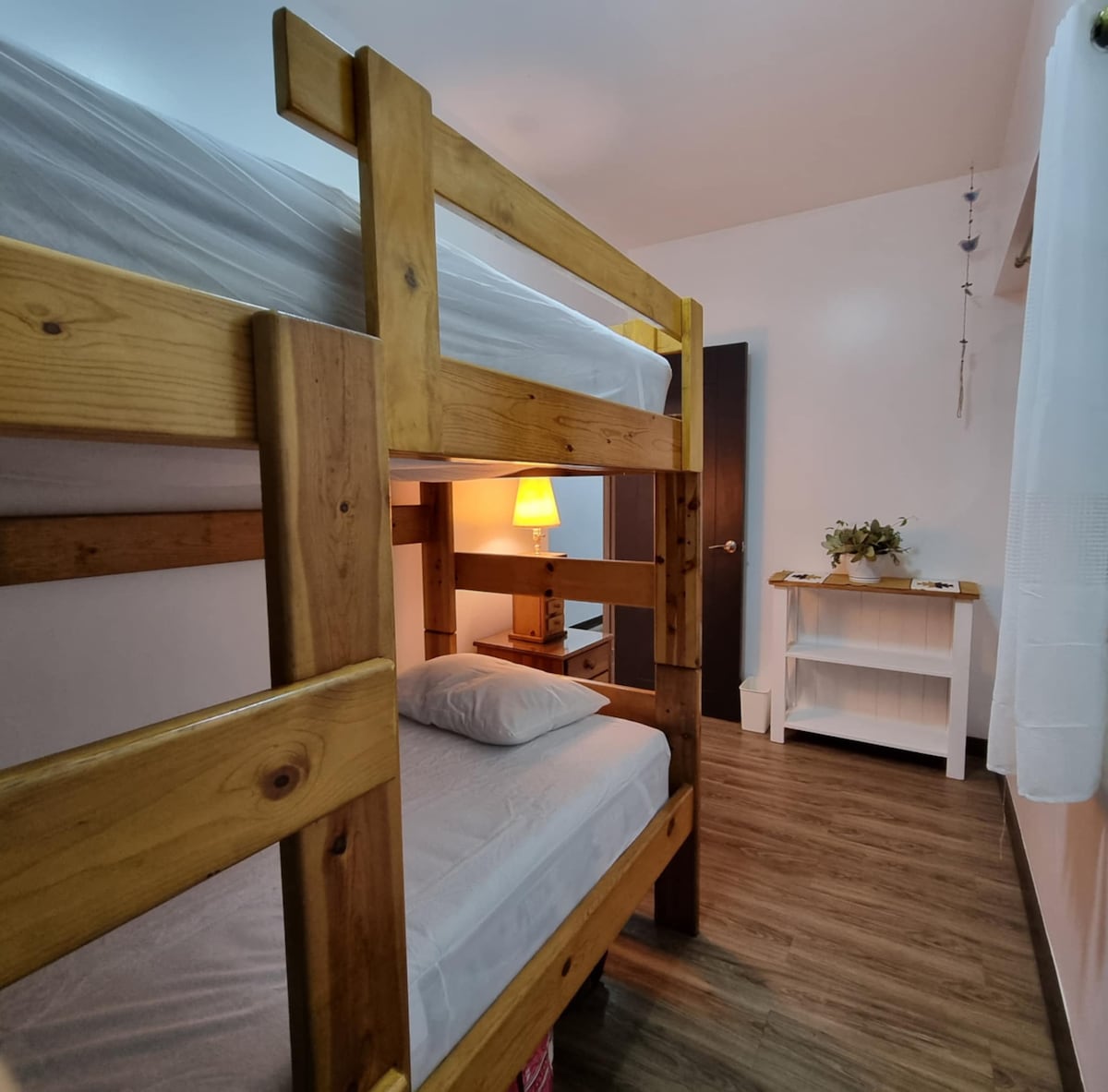 A wooden bunk bed is situated in a softly lit room, with a bedside lamp illuminating a small nightstand. A white shelf is visible against the wall, adding functional storage. The warm wood tones complement the neutral wall colors and enhance the cozy environment.