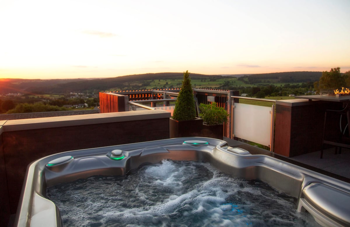 A hot tub with bubbling water is positioned on a terrace, offering expansive views of rolling hills under a sunset sky. Natural greenery is present in potted plants nearby, enhancing the serene outdoor space.