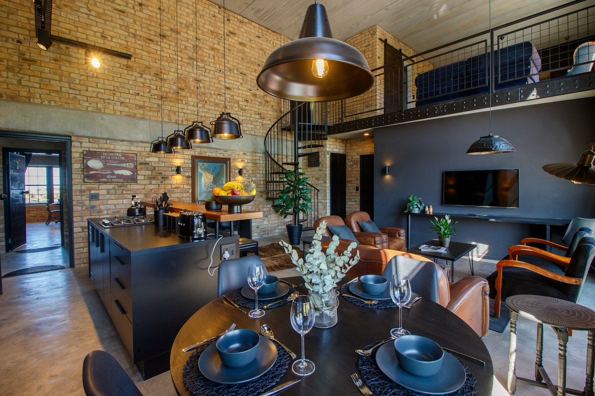 An open-plan kitchen, dining, and living area features exposed brick walls and large windows that allow natural light. A circular dining table is set for meals, while a cozy seating arrangement invites relaxation. A spiral staircase leads to the mezzanine sleeping area above.