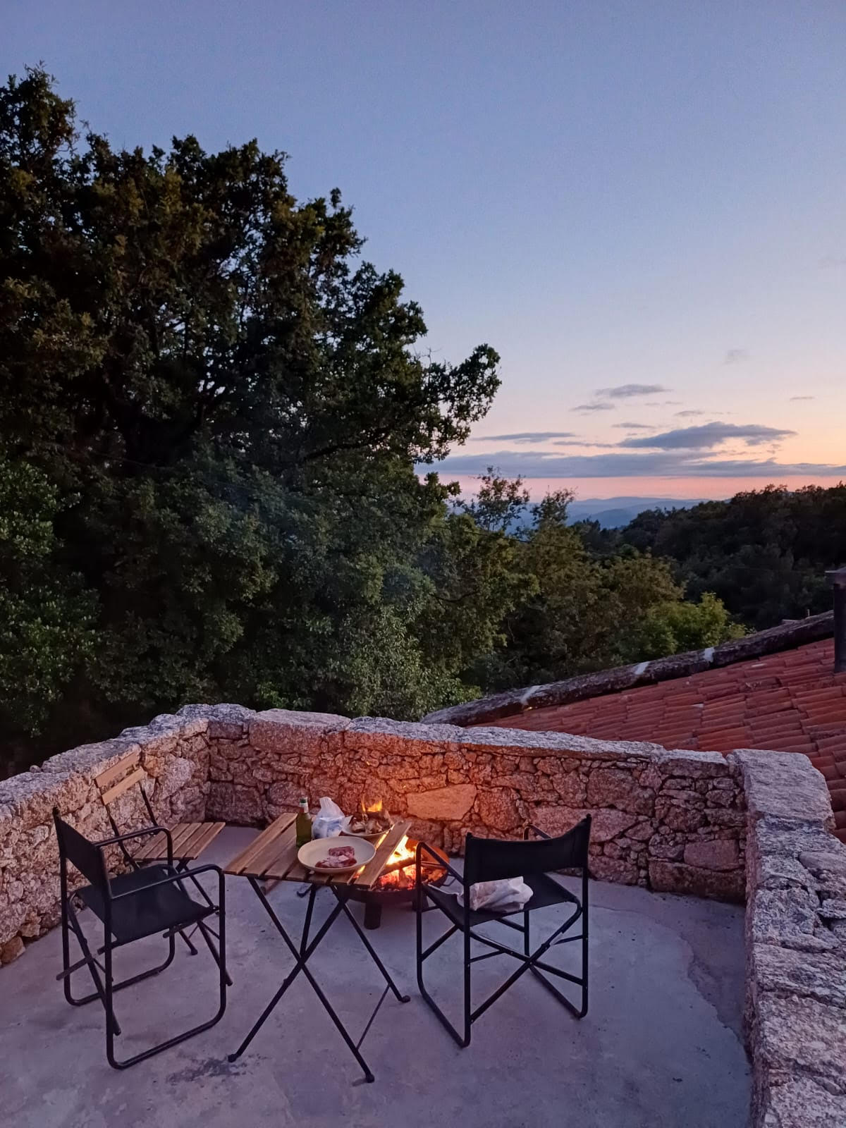 A stone terrace is adorned with two black chairs and a small round table, set for an evening gathering. Soft lights from a fire pit glow warmly in the center, with greenery from surrounding trees creating a serene backdrop against the twilight sky.