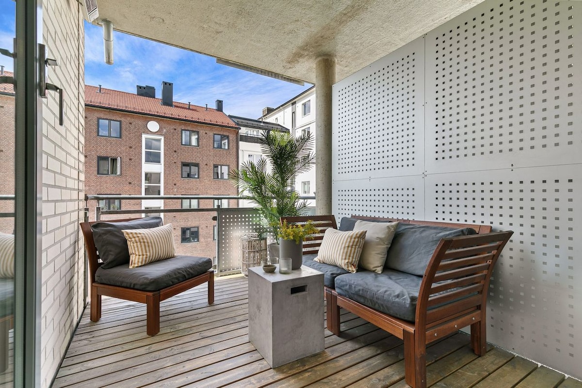 A private balcony features wooden decking and modern seating arrangements. Two cushioned chairs face a small concrete table. Decorative elements include potted greenery. The balcony is enclosed with perforated panels, offering a sense of privacy while overlooking adjacent buildings.