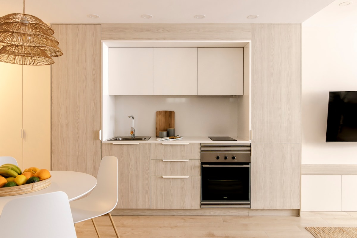 The kitchen area features modern cabinetry in light wood tones, with a compact cooking space including an electric stove and oven. A small sink is situated beside a wooden cutting board. A round dining table with white chairs is positioned nearby, accented by a fruit bowl.