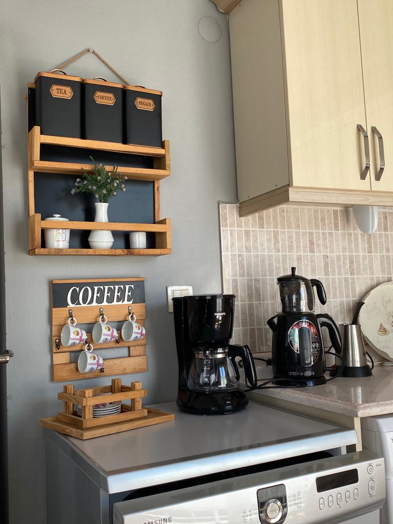 A well-organized kitchen corner features wooden shelves displaying labeled containers for tea, sugar, and snacks. A coffee station showcases multiple coffee-making appliances, including a coffee maker and a kettle. Decorative cups hang below a sign reading 'COFFEE,' contributing to a functional yet inviting space.