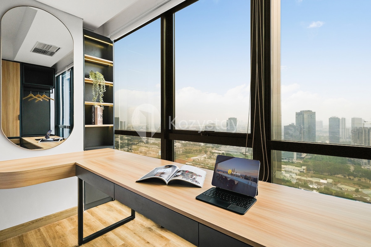 A spacious workspace is provided with a wooden desk positioned beside large windows offering panoramic city views. A laptop rests on the desk next to an open magazine, while a round mirror reflects the natural light streaming into the room.